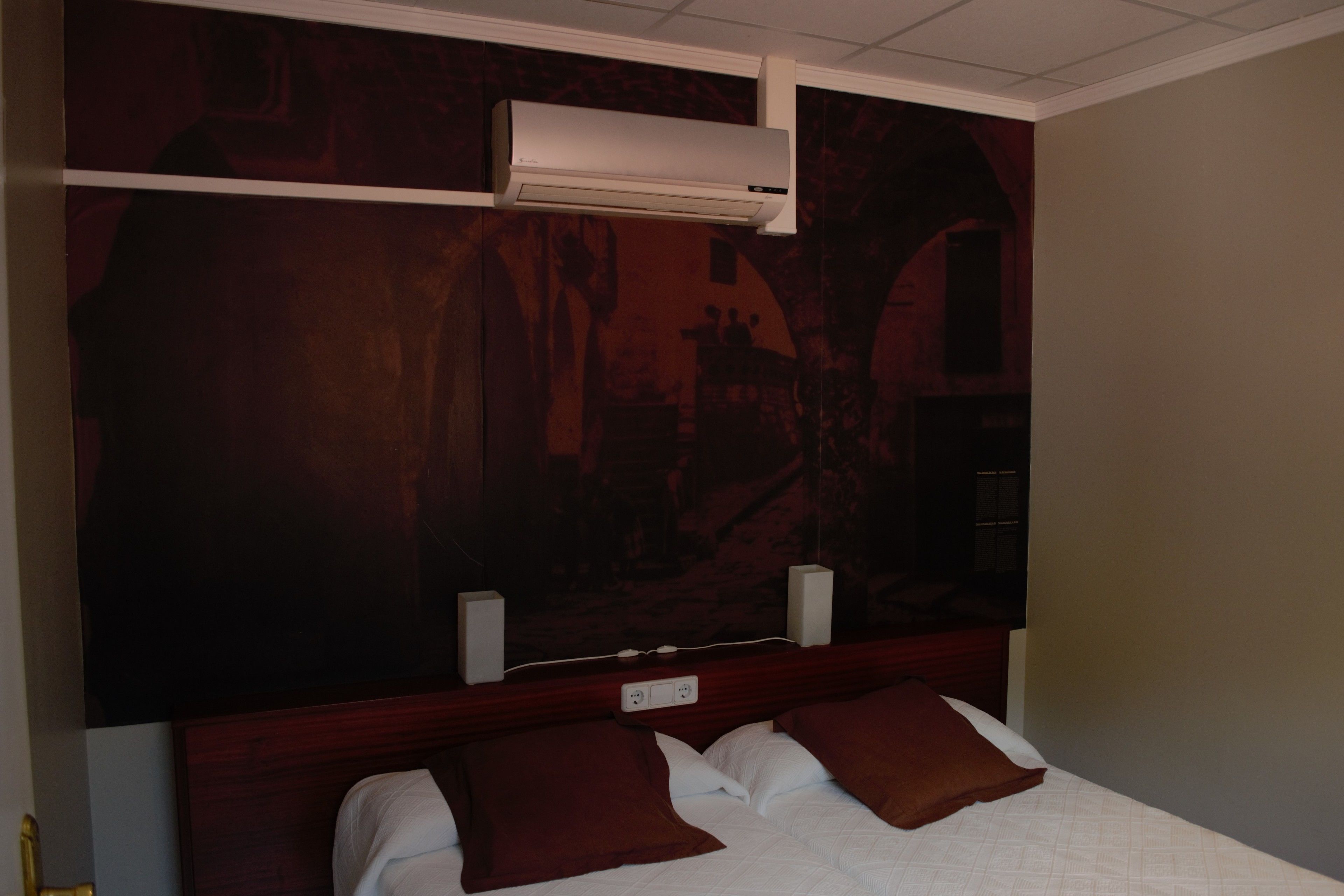 A hotel room with a large bed, two brown pillows, a white comforter, a wooden headboard, and a wall-mounted air conditioner.
