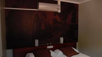 A hotel room with a large bed, two brown pillows, a white comforter, a wooden headboard, and a wall-mounted air conditioner.