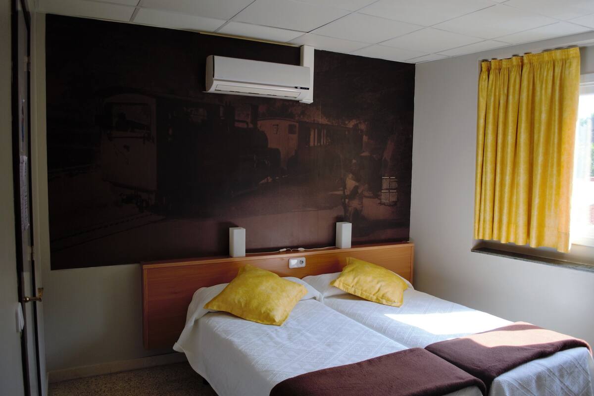 Una habitación de hotel con una cama, dos almohadas, un cabecero, un equipo de aire acondicionado, un ventanal con cortinas amarillas y una obra de arte colgada en la pared.