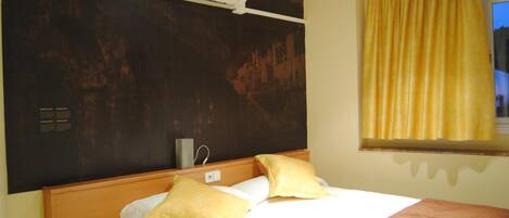 A hotel room with a bed, a bedside table with a remote control, a wall-mounted air conditioner, and a window with yellow curtains.