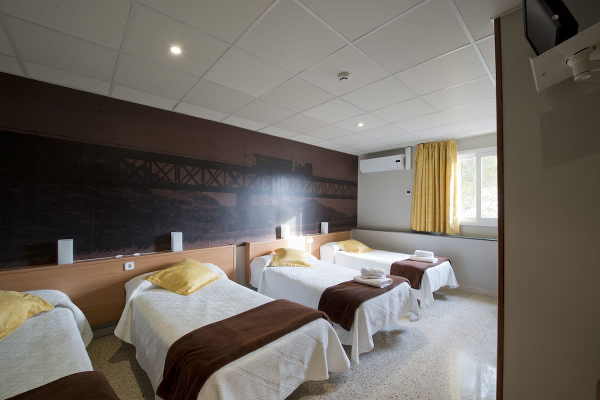 Habitación de hotel con cuatro camas, un mural grande en la pared, una ventana con cortinas y un equipo de aire acondicionado.