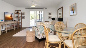 Ett rymligt vardagsrum med ett matbord med glasskiva omgivet av flätade stolar, en soffa med blommigt tyg och en tv-apparat placerad på ett träställ.