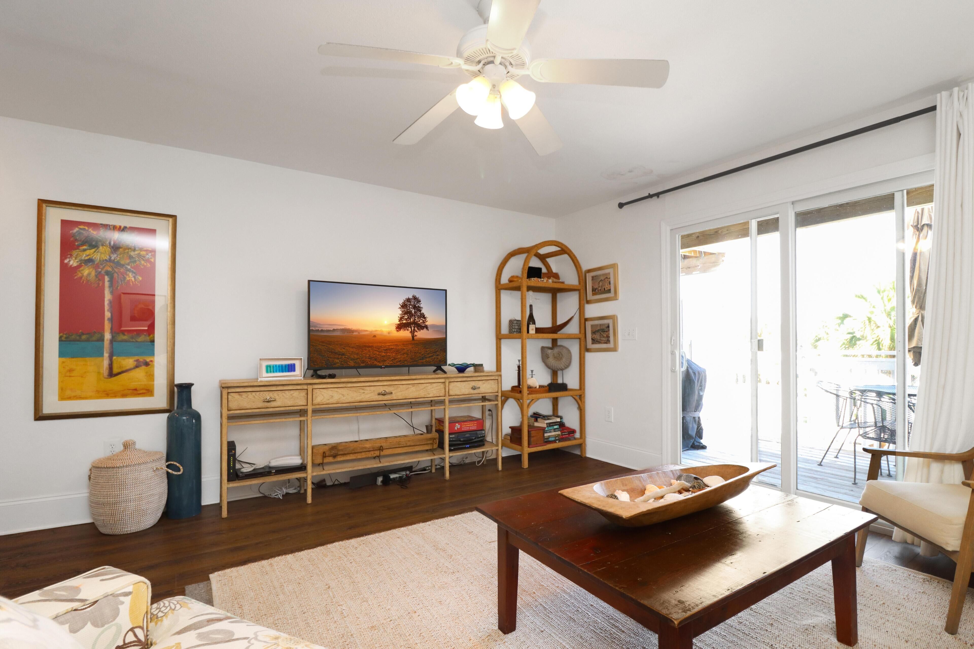 A living room with a wooden coffee table, a television on a wooden stand, a large window with curtains, and a ceiling fan.
