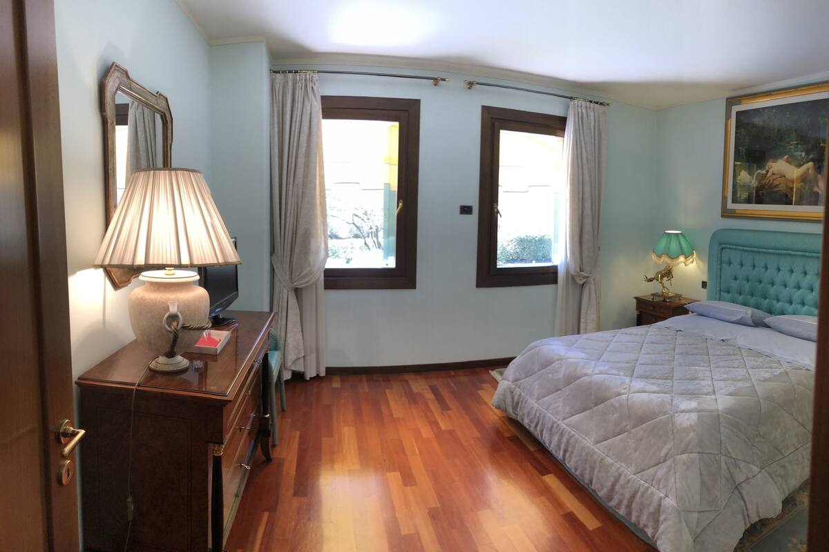 Une chambre à coucher comprenant un lit, une table de chevet, une lampe, une commode, un miroir et deux fenêtres avec des rideaux.