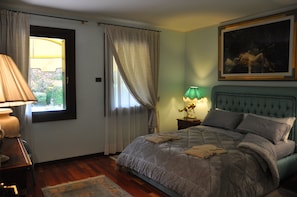 A bedroom with a bed, a nightstand, a lamp, a window with curtains, and a painting on the wall.