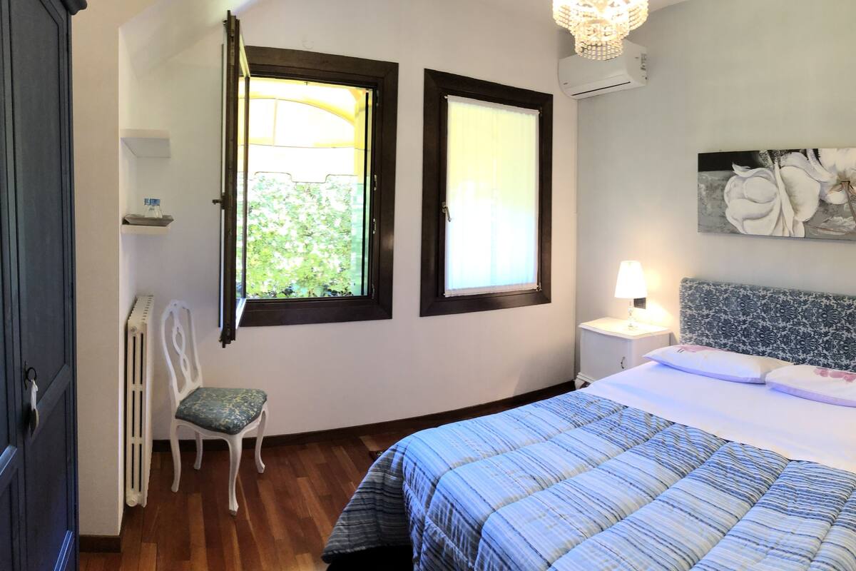 Une chambre à coucher avec un lit, deux lampes de chevet, une table de chevet, une chaise, une fenêtre et un tableau accroché au mur.