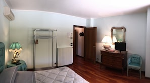 A hotel room with a bed, a desk with a lamp, a dresser, a chair, a radiator, and a door leading to another room.