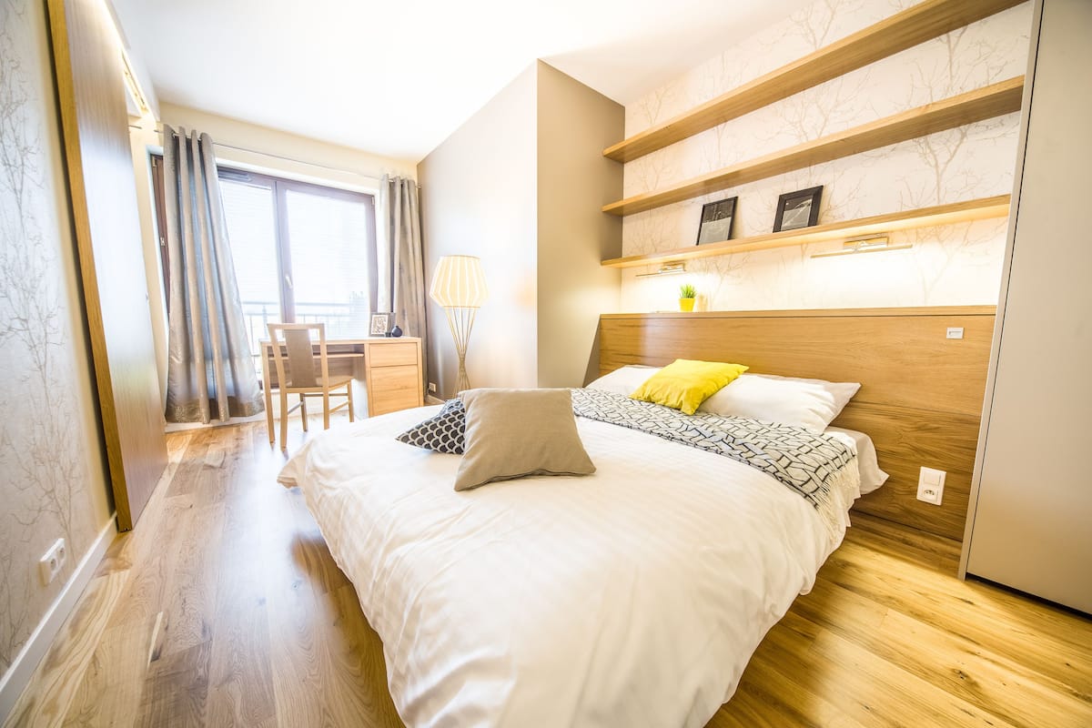 Une chambre à coucher avec un grand lit, un bureau avec une chaise, une fenêtre avec des rideaux et des étagères intégrées ornées d’objets décoratifs.