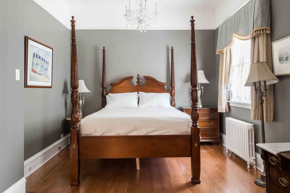 A four-poster bed with a wooden headboard, two bedside tables with lamps, a radiator, and a chandelier.