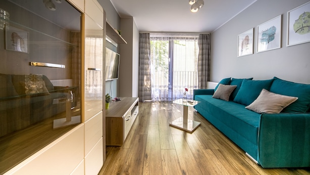 Een moderne woonkamer met een blauwgroene bank, houten vloer, een glazen salontafel en een groot raam met gordijnen.