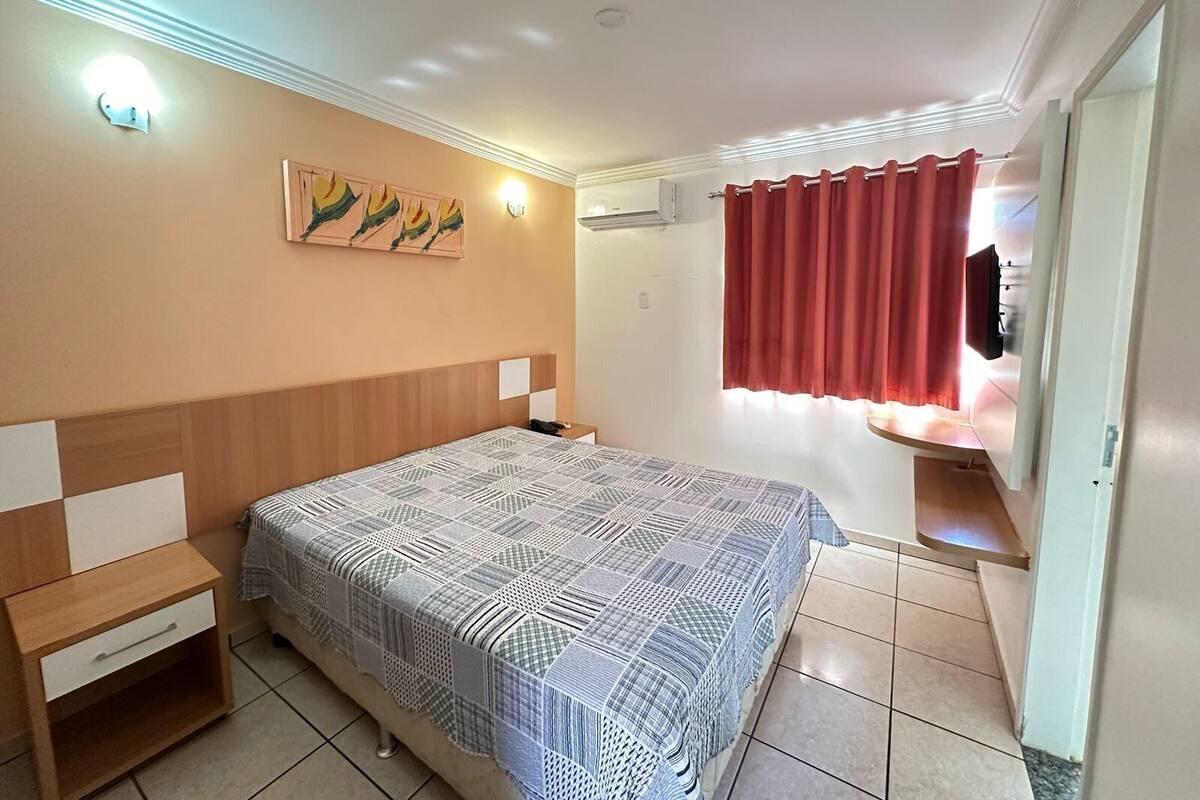 A bedroom with a bed, a nightstand, a wall-mounted shelf, a window with curtains, and a painting on the wall.