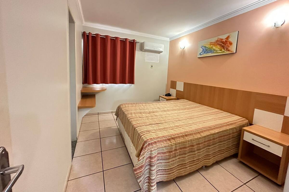 A hotel room with a bed, a nightstand, a small table, a red curtain, a wall-mounted air conditioner, and a painting.