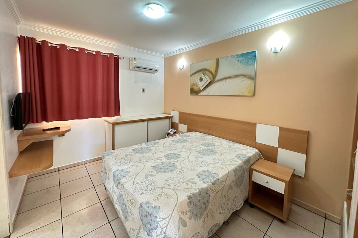 A hotel room with a bed, bedside tables, a desk, a chair, a TV, a painting, and an air conditioning unit.