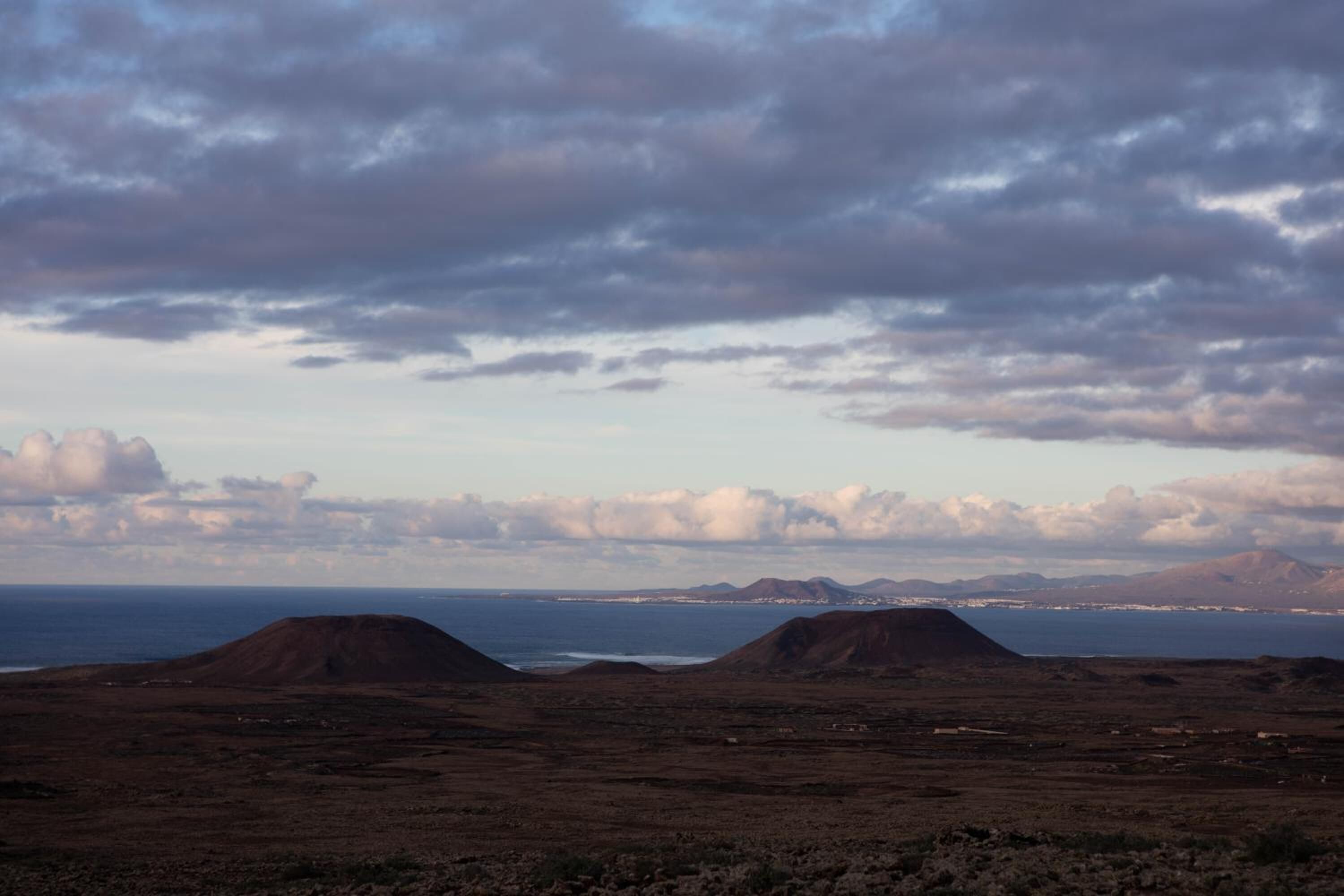 A barren landscape with two prominent hills and a distant coastline under a cloudy sky.