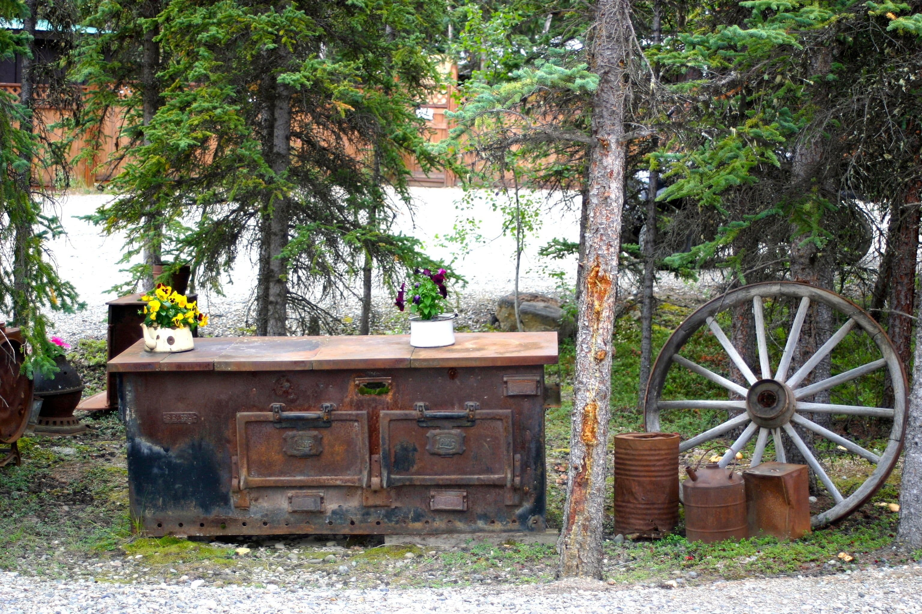 A rustic wooden bench with a planter, surrounded by trees and a large wooden wheel.