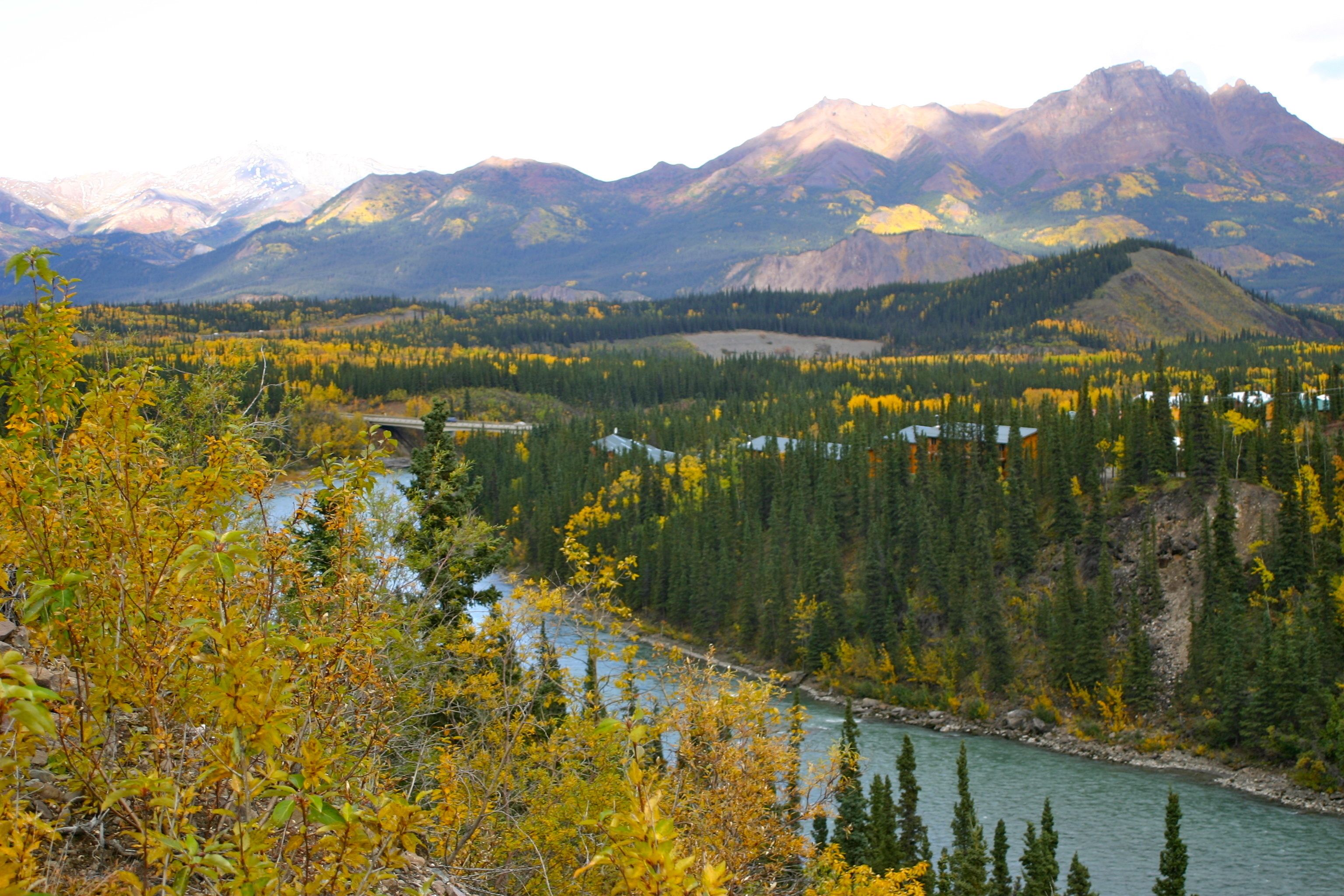 A mountainous landscape with a river, dense forests, and a few buildings in the distance.