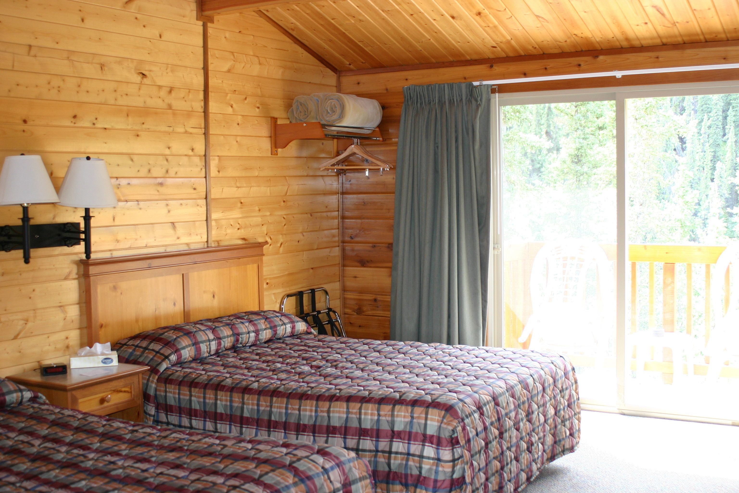 Habitación de cabaña de madera con dos camas, una mesita de noche, una lámpara, un perchero y un ventanal con vistas a una terraza.