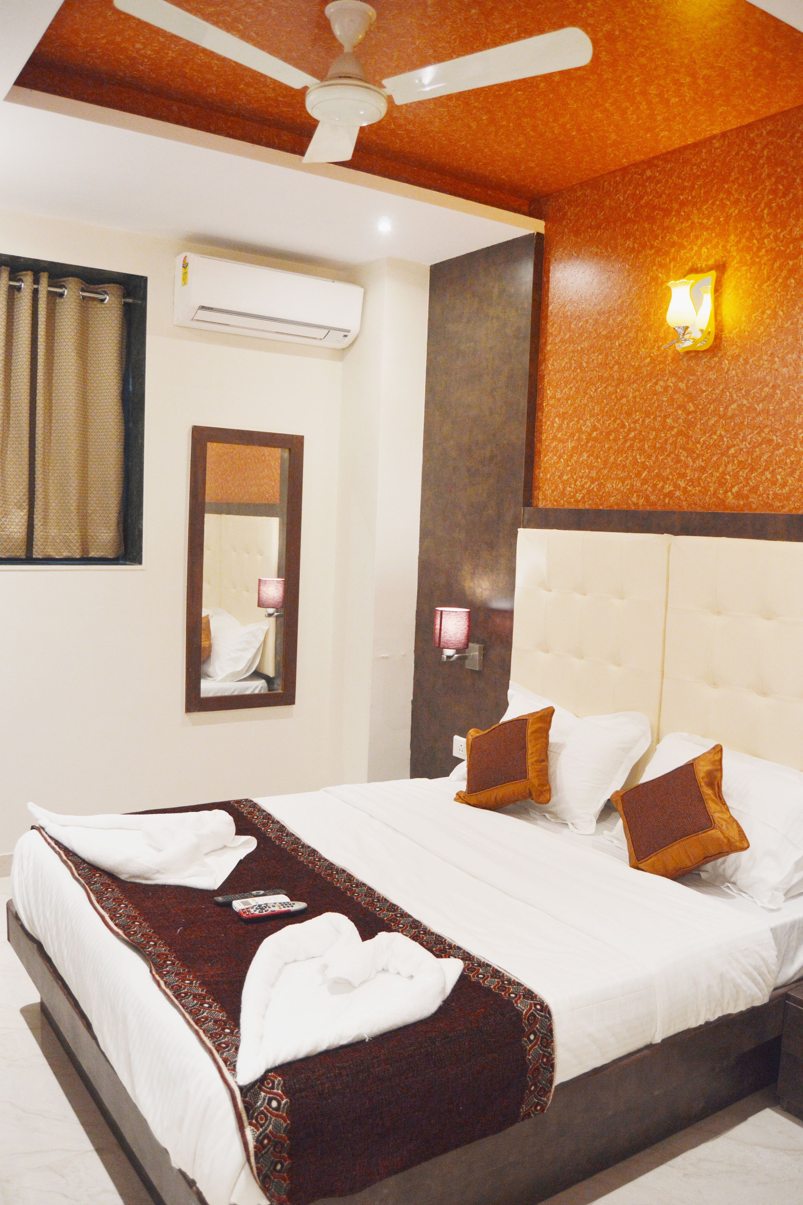 A hotel room with a bed, a ceiling fan, an air conditioning unit, a mirror, and a wall-mounted light fixture.