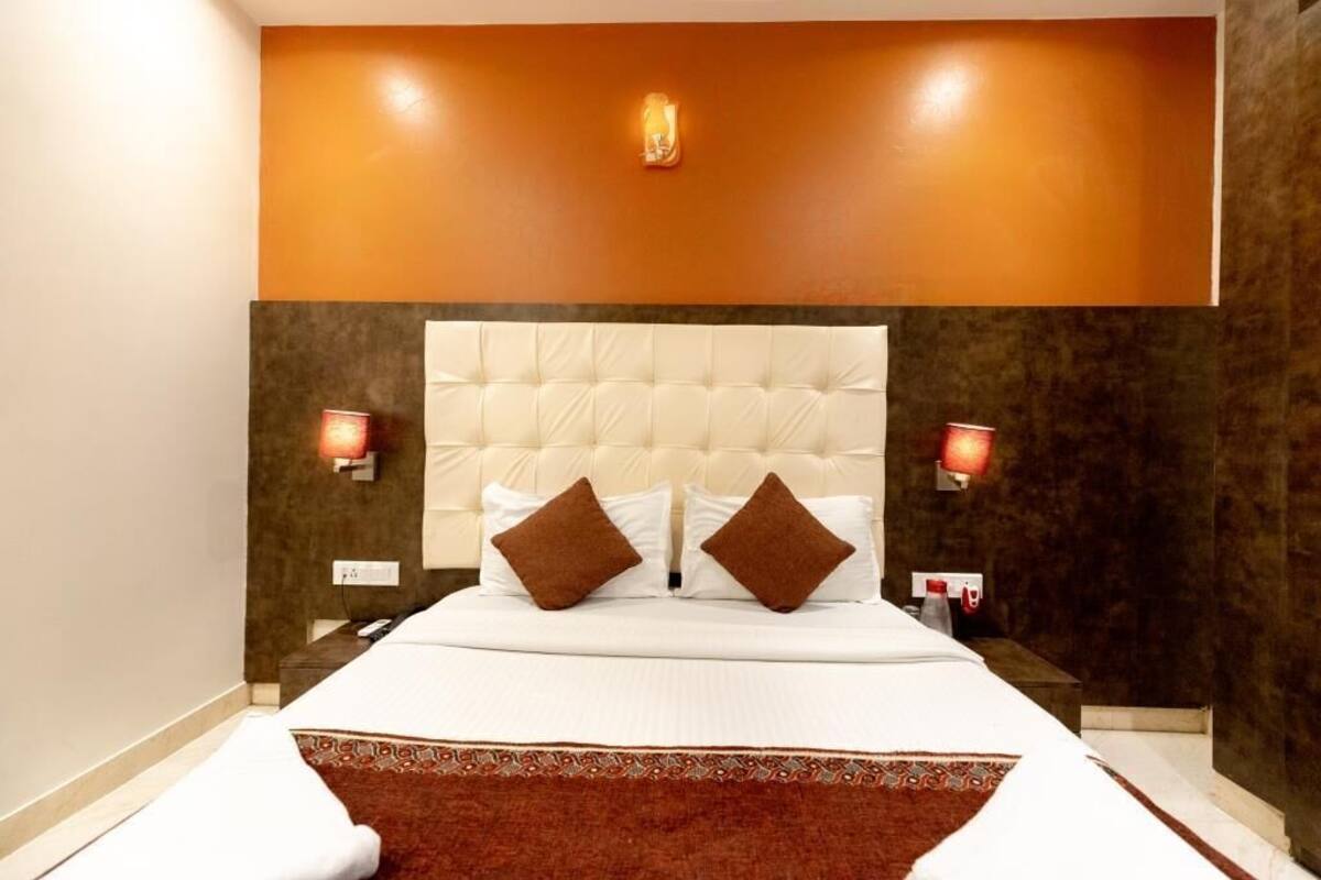 A hotel room with a tufted headboard, two pillows, a bedside table with a lamp, and a patterned rug.