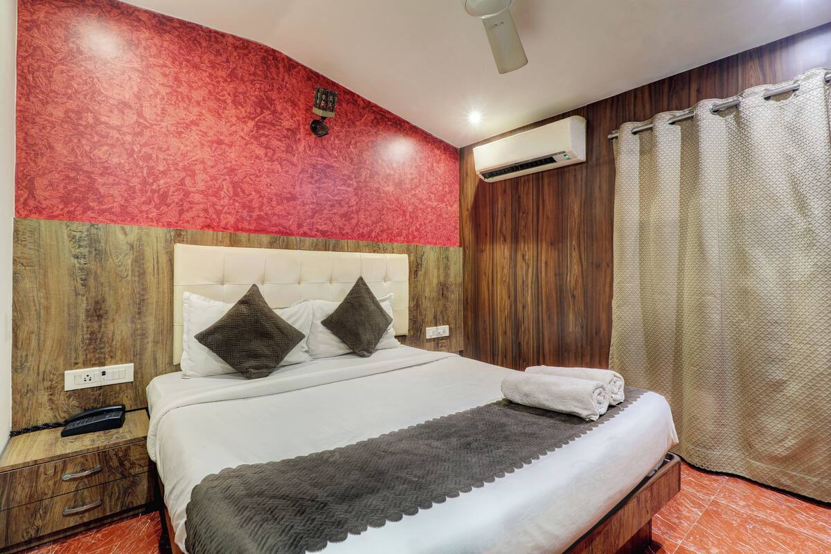A hotel room with a wooden headboard, a bed with white linens, two pillows, a remote control on the nightstand, and an air conditioning unit on the wall.