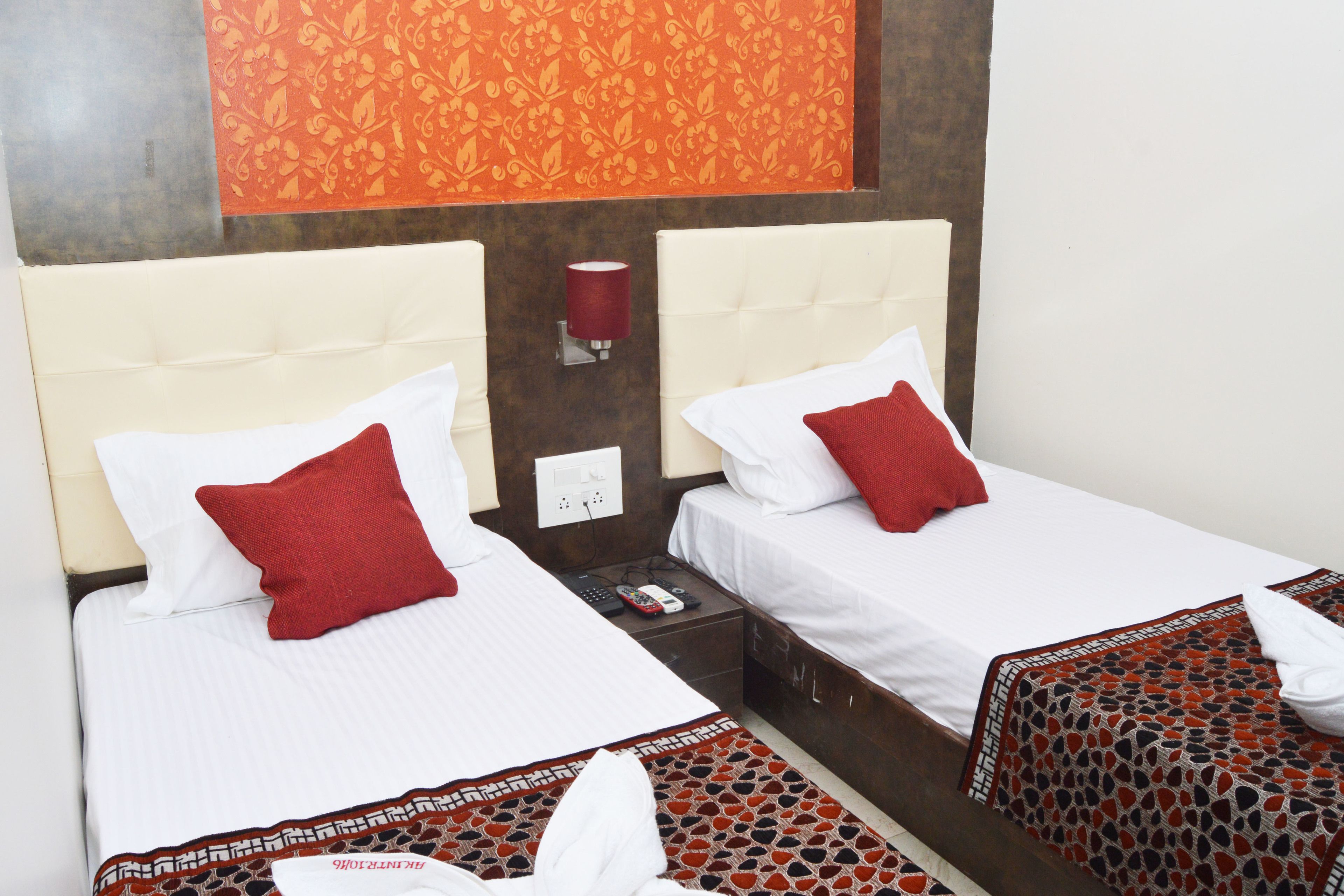 A hotel room with two beds, each with white linens and red pillows. There is a patterned rug on the floor and a wall-mounted lamp between the beds.