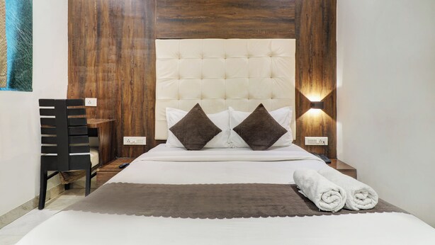 A bedroom with a tufted headboard, two bedside lamps, a bed with white bedding, a brown throw blanket, and a rolled towel on the bed. - Hotel AK International (Mumbai)