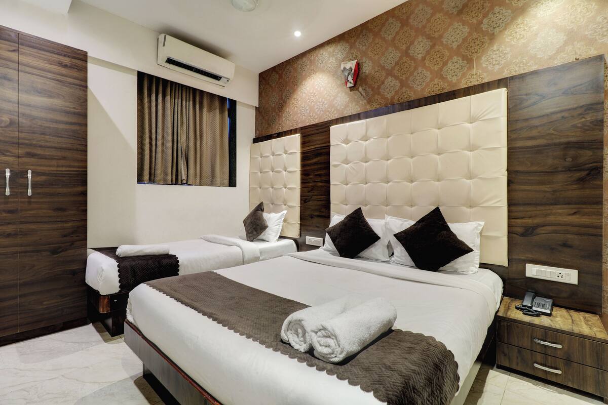 A hotel room with a large bed, two smaller beds, a wooden wardrobe, a bedside table with a phone, and an air conditioning unit.