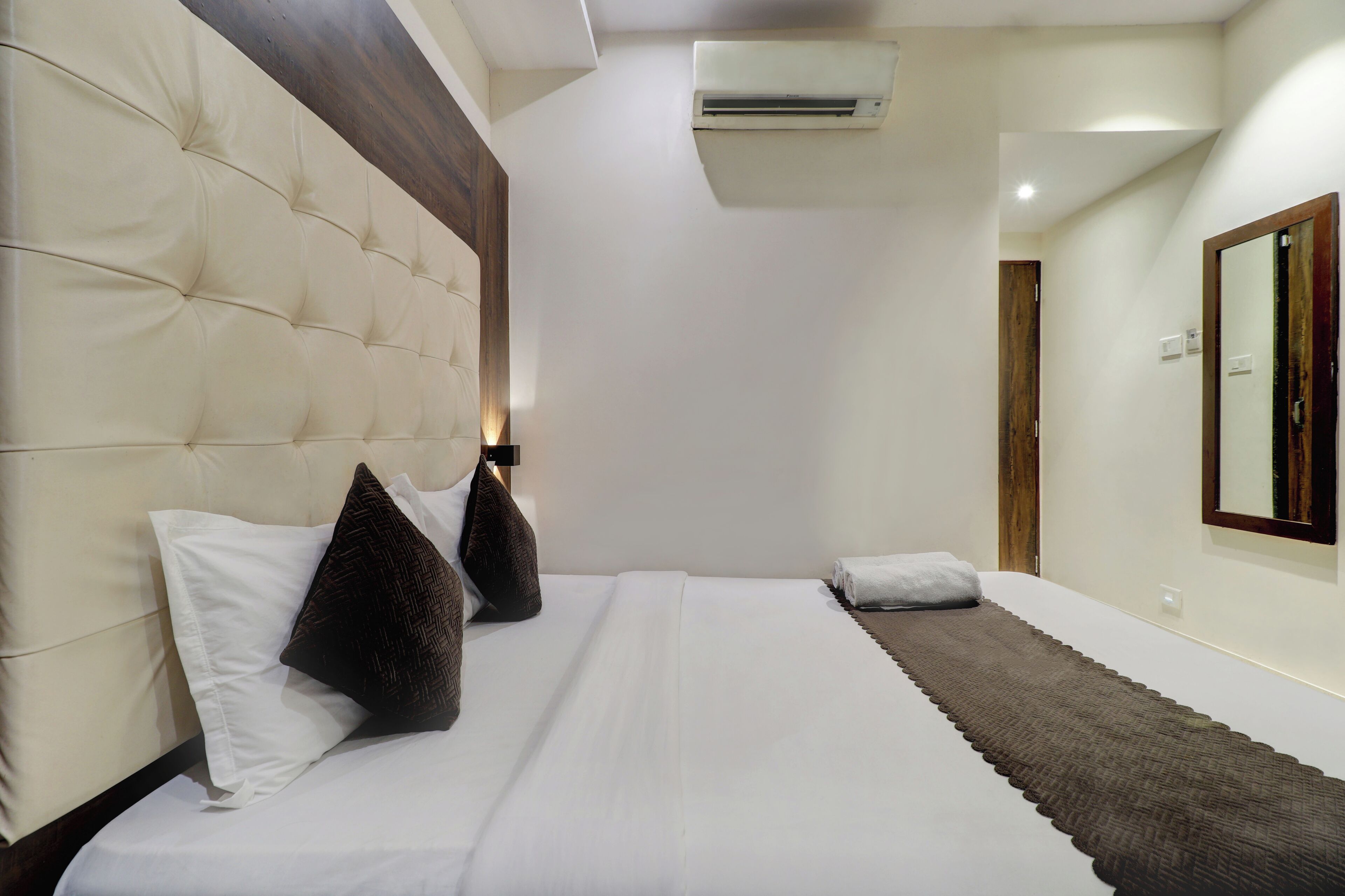 A bedroom with a tufted headboard, a large mirror, and a neatly made bed with white linens and brown accents.