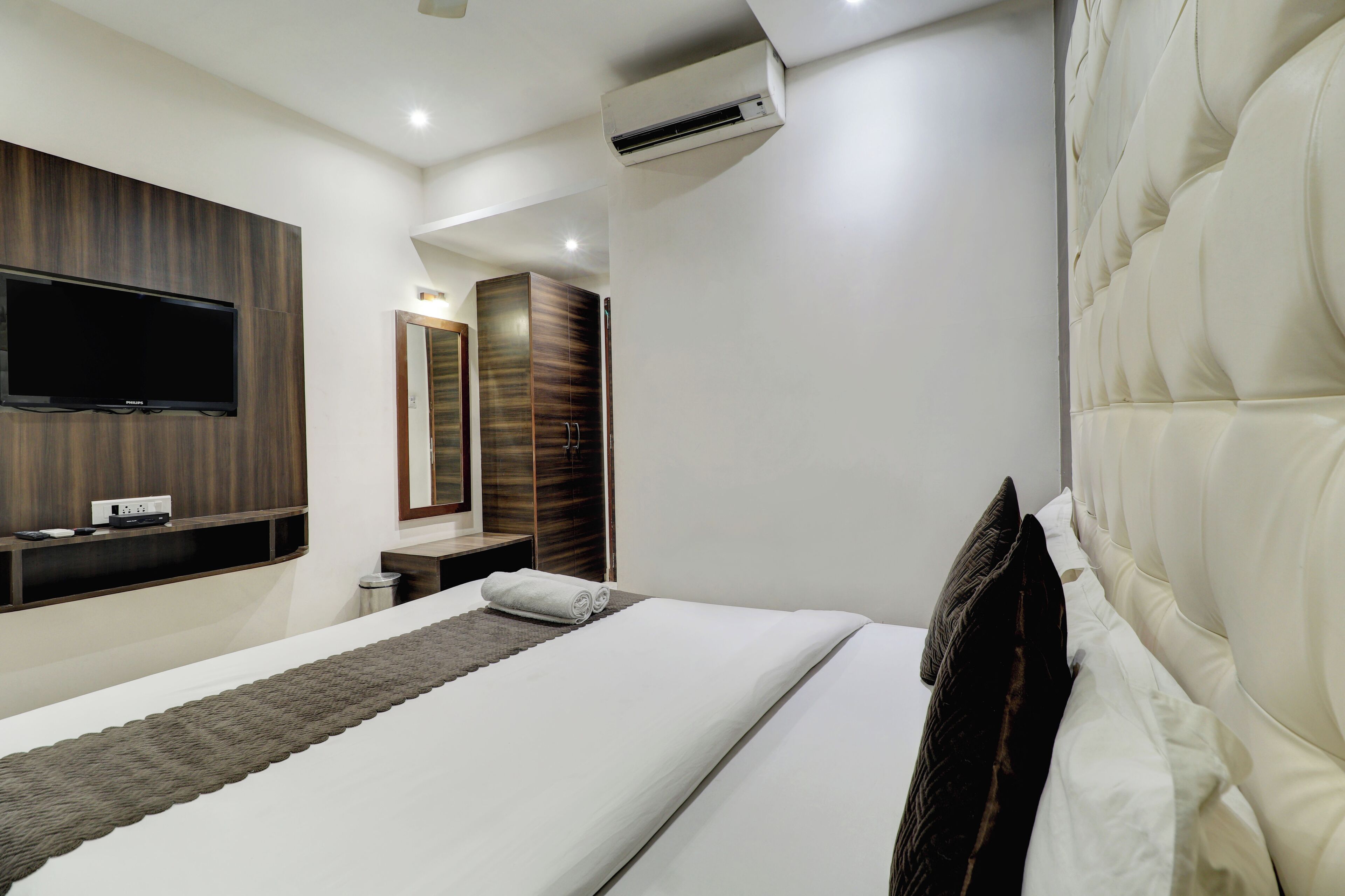 A modern hotel room with a large bed, a flat-screen TV mounted on the wall, a vanity with a mirror, and a built-in air conditioning unit.