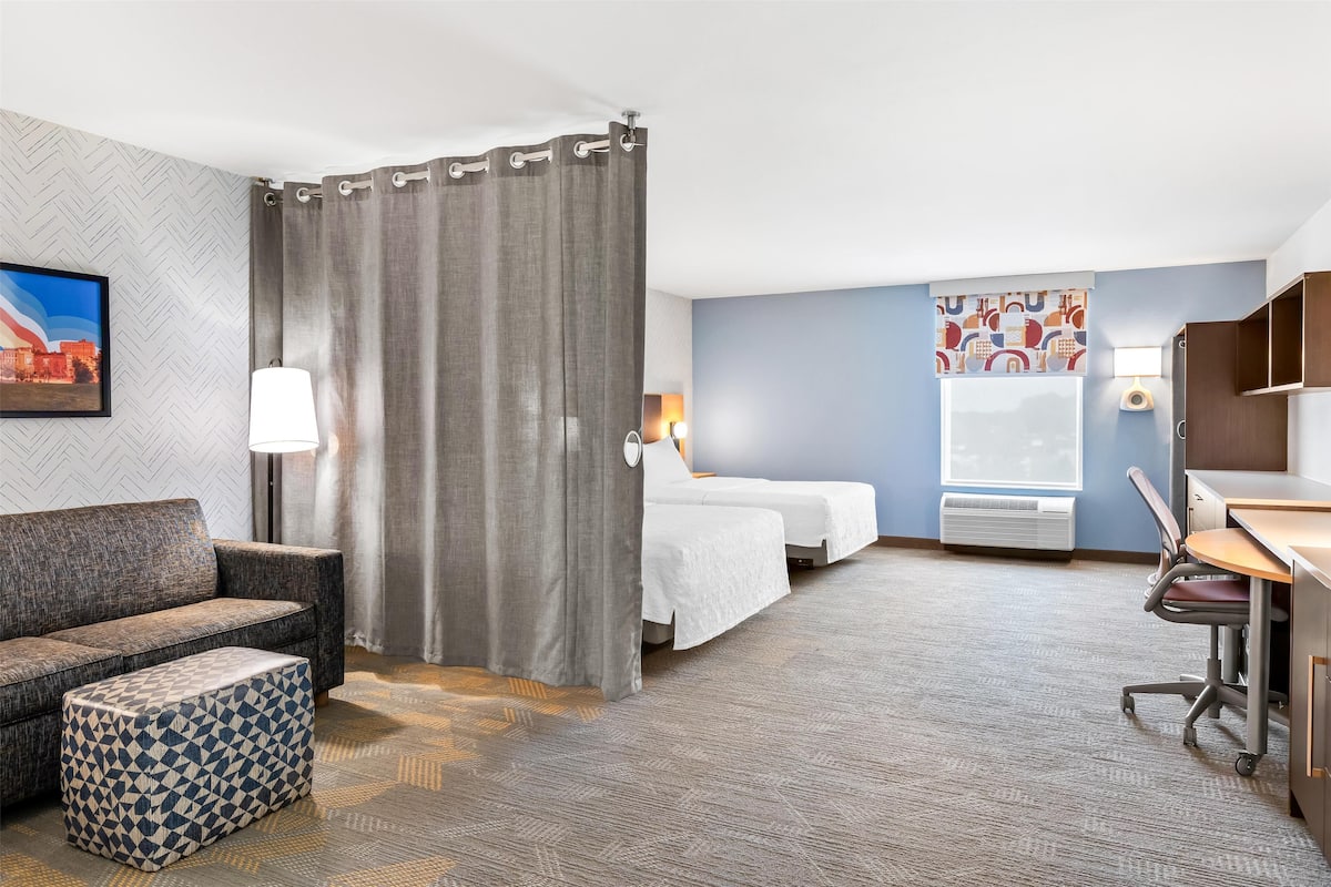 Ein Hotelzimmer mit einer Couch, zwei Betten, einem Schreibtisch, einem Stuhl, einem gemusterten Hocker, einem Vorhang, einer Wandleuchte, einem Fenster mit Aussicht und einem Gemälde.
