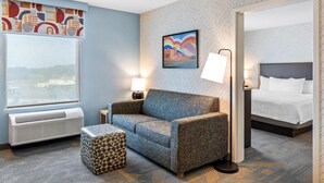 A hotel room with a sofa, ottoman, and a bed. There is a window, a wall-mounted picture, and a lamp on a bedside table.