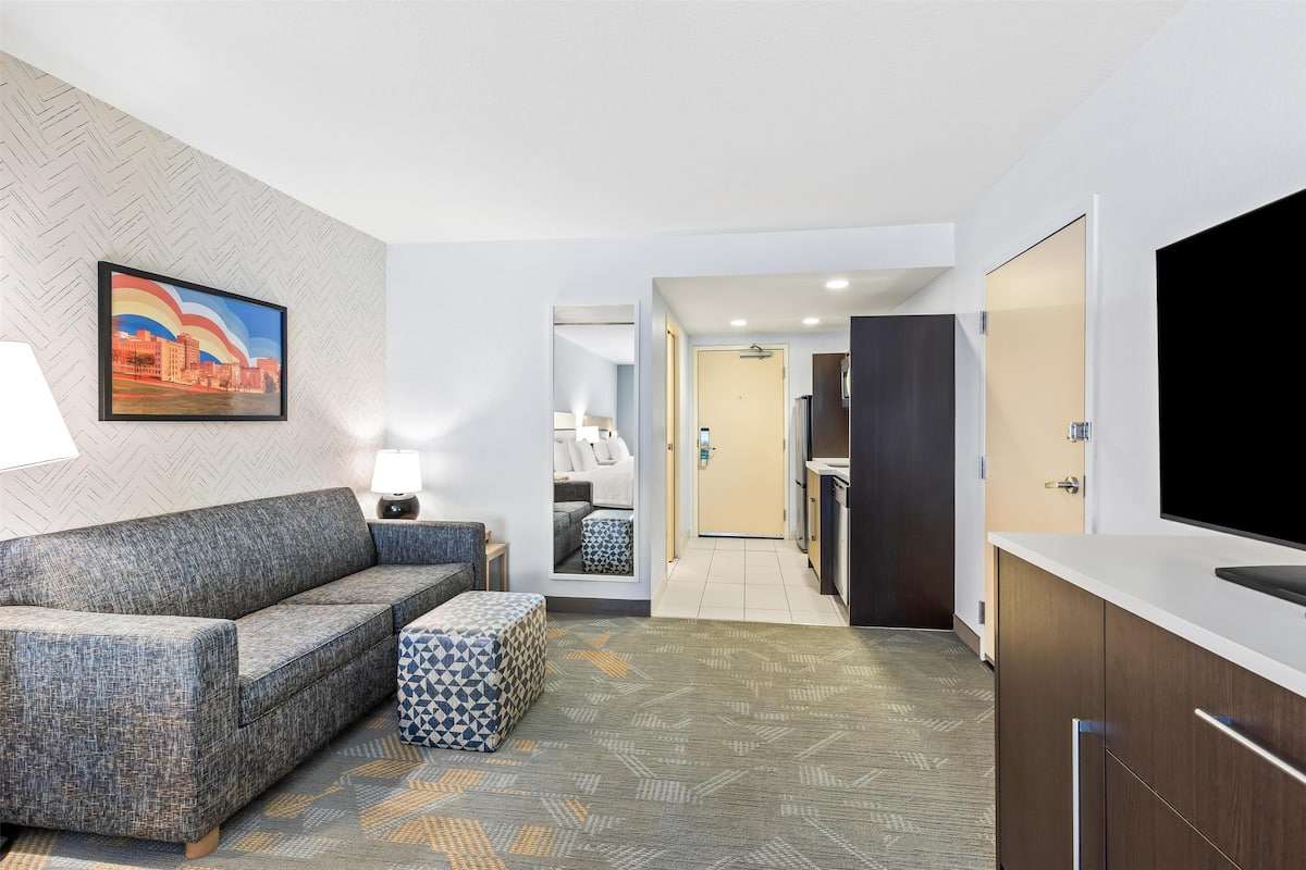 Ein Hotelzimmer mit Sofa, Hocker und Fernseher. Ein Flur führt zu einem Badezimmer und einer Küchenzeile.