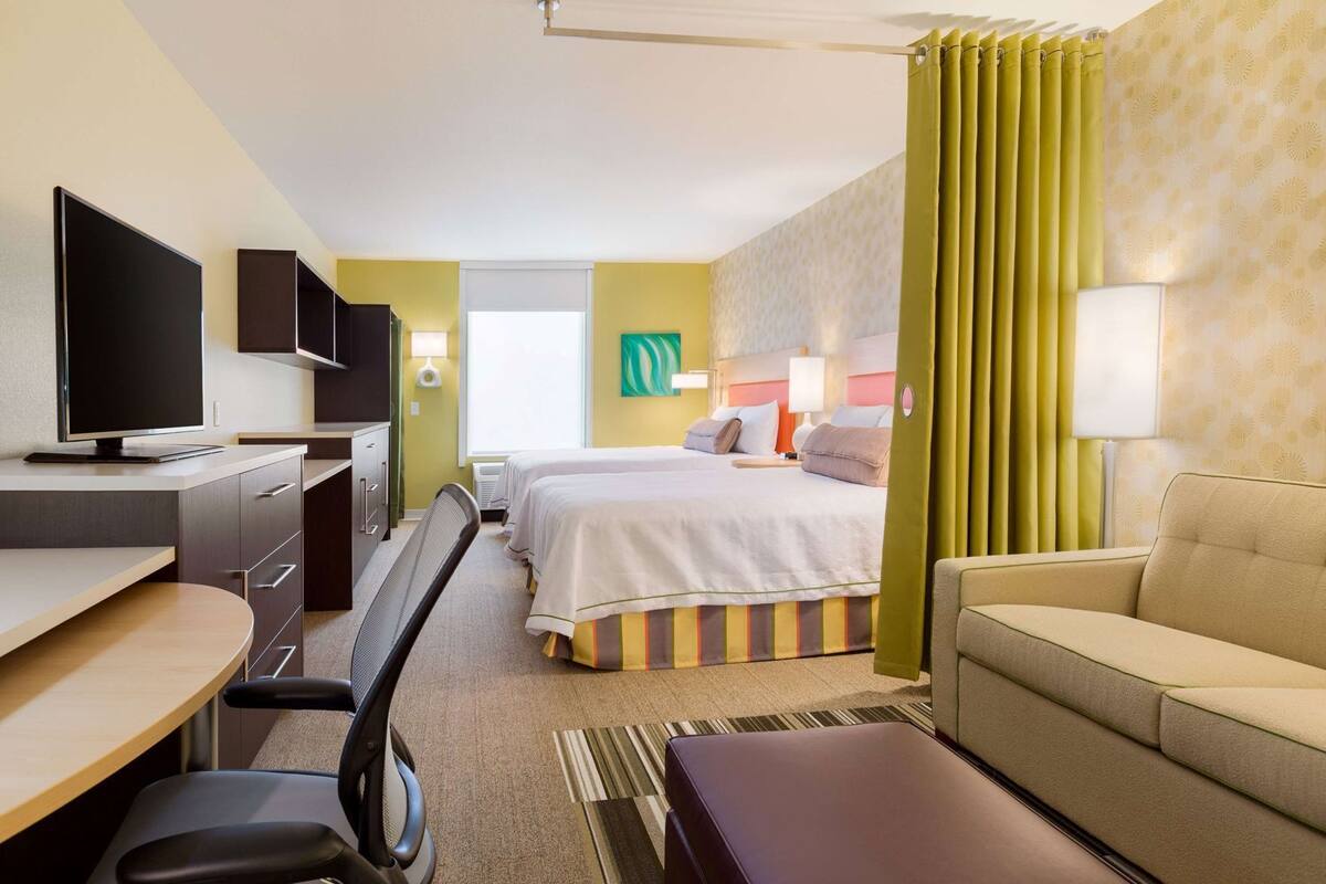 Quarto de hotel com escrivaninha, duas camas, sofá, televisão e janela com cortinas.