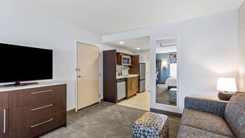 Une chambre d’hôtel moderne équipée d’un téléviseur à écran plat, d’un canapé, d’un pouf à motifs, d’un petit coin cuisine et d’un miroir qui reflète la pièce.