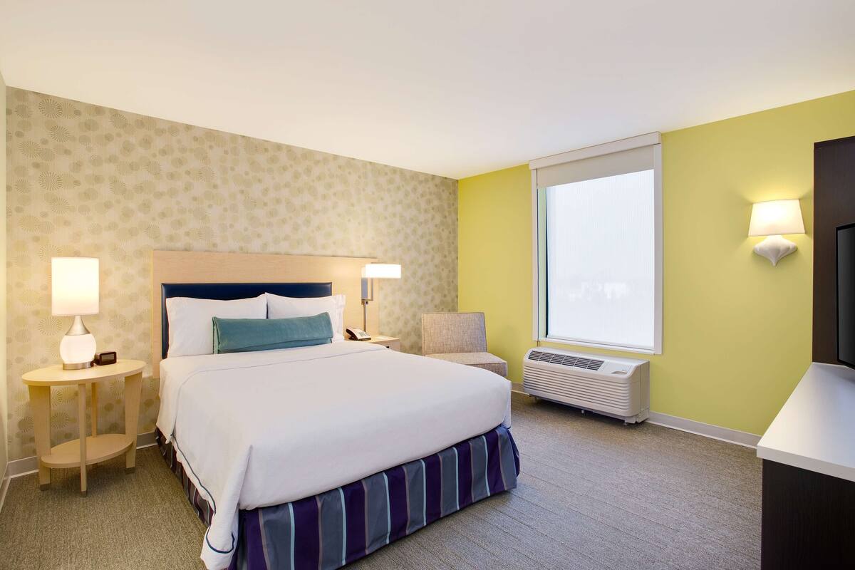 Quarto de hotel com uma cama grande, duas mesas de cabeceira com luminárias, uma cadeira, janela com vista e ar condicionado.