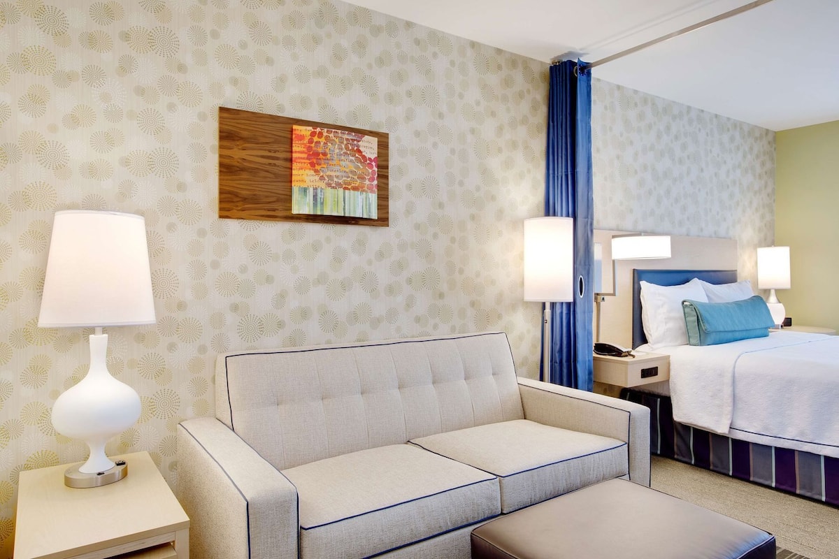 Quarto de hotel com sofá, cama, mesa de cabeceira, luminária e um quadro na parede.