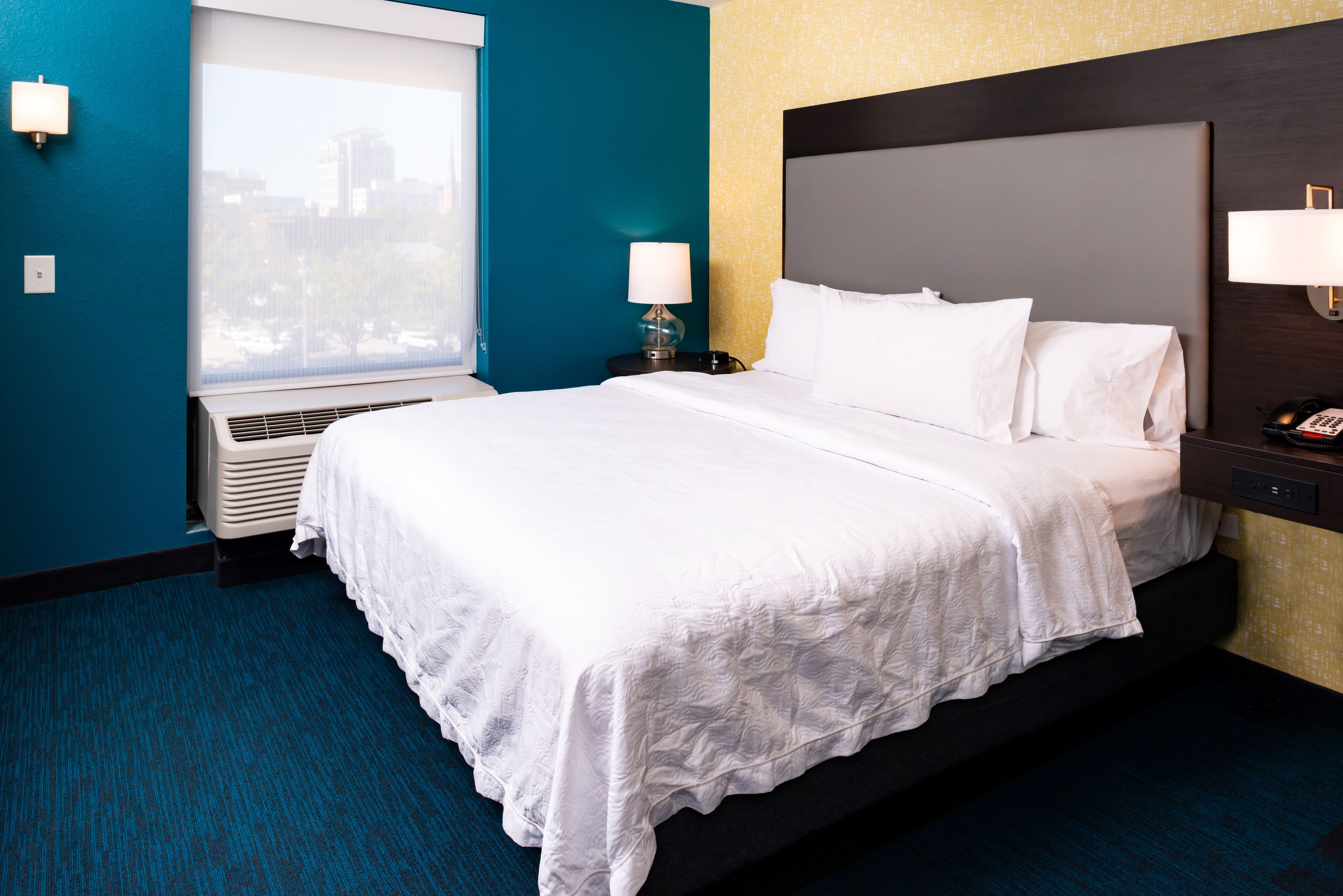 Habitación de hotel con una cama grande, una ventana con vistas a edificios, aire acondicionado de pared y una mesita de noche con lámpara.