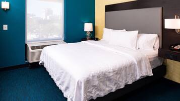 Habitación de hotel con una cama grande, una ventana con vistas a edificios, aire acondicionado de pared y una mesita de noche con lámpara.