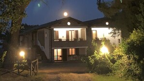 Une maison à deux étages avec un escalier apparent menant à l’étage supérieur, entourée d’arbres et éclairée par des lampes extérieures.