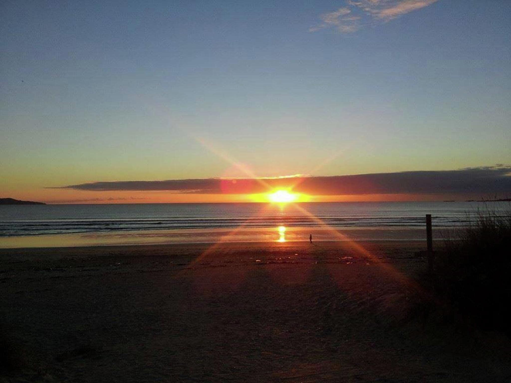 A beach at sunset with the sun low on the horizon, creating a reflection on the water.