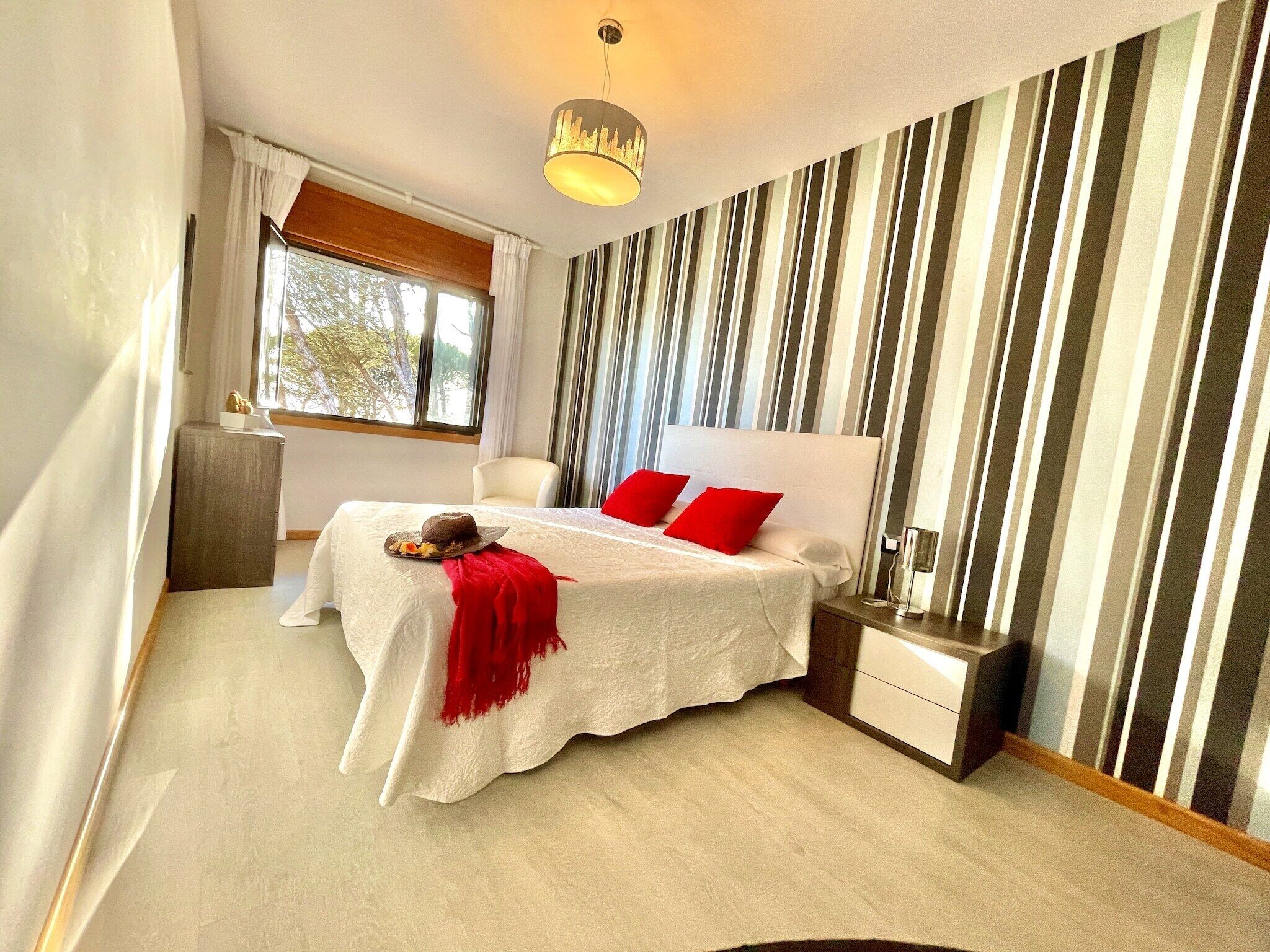 A bedroom with a bed, a nightstand, a lamp, a window, and a striped wall.