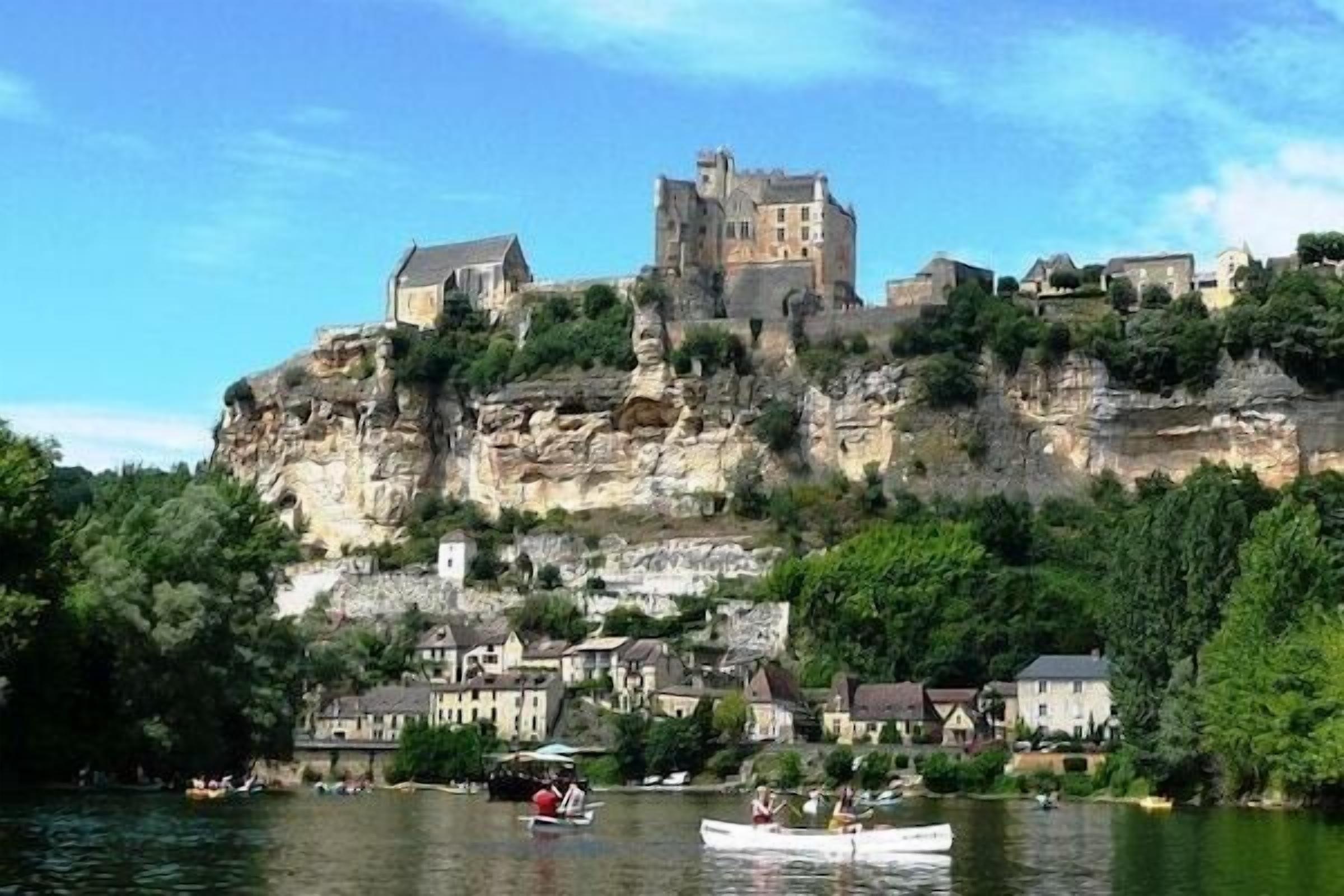 Un castello a picco su una scogliera che si affaccia su un fiume con barche, circondato da case.