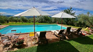 Une piscine entourée de verdure, avec des chaises longues et des parasols, sous un ciel dégagé.