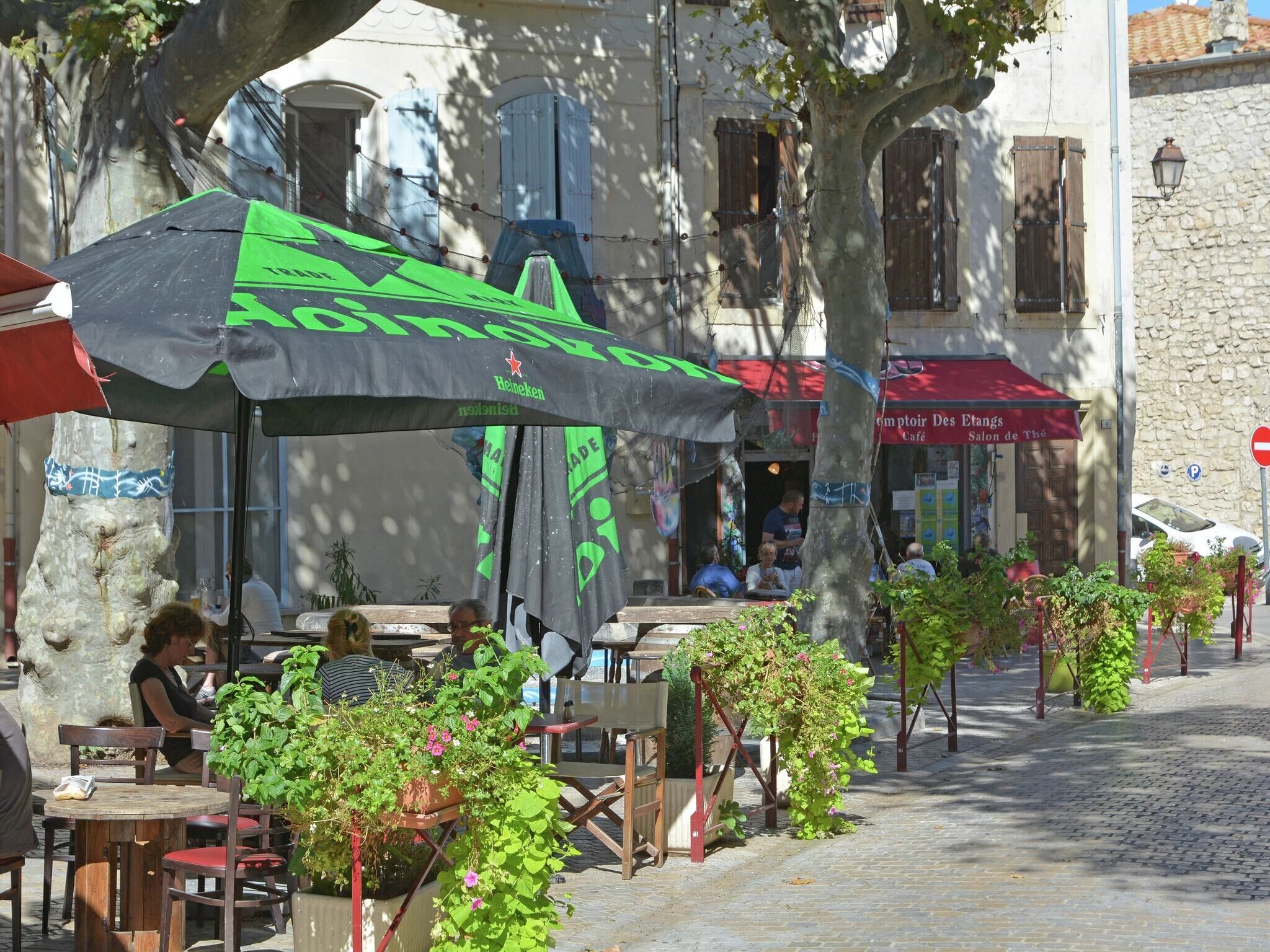Des tables et des chaises sont disposées à l’extérieur, un café avec un auvent rouge se trouve sur place, et la rue est pavée.