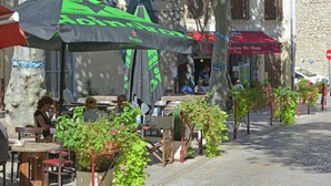 Des tables et des chaises sont disposées à l’extérieur, un café avec un auvent rouge se trouve sur place, et la rue est pavée.