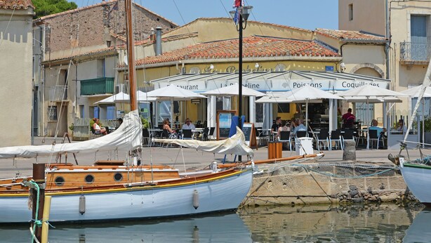 Un restaurant situé au bord de l’eau, avec une terrasse et un voilier amarré à proximité.