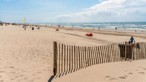 Platja sorrenca amb una tanca de fusta, una bandera groga i persones a la distància.