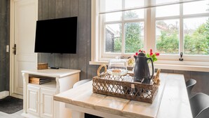 Una habitació amb televisió penjada a la paret, una finestra amb vista a vegetació, un armari blanc i una taula amb una cistella i una gerra.