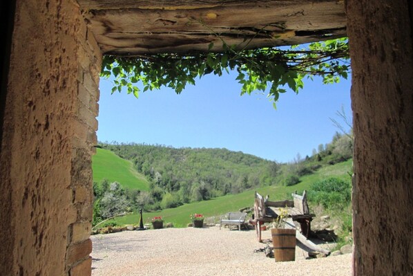 A rustic stone archway leading to a gravel area with a bench and potted plants, overlooking a green hillside.