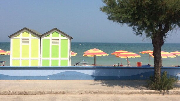 A beach with two green and white beach huts, a row of red and yellow umbrellas, and a tree in the foreground. - Cozy Couples Getaway w/ wifi (Pergola)